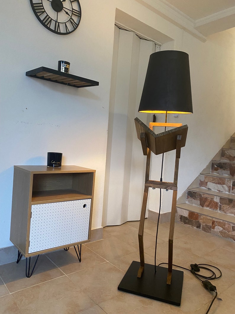 May include: A wooden floor lamp with a black lampshade. The lamp is shaped like a person with a long neck and legs. The lamp is standing on a black base.