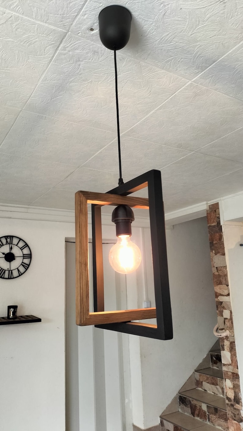 May include: A modern pendant light fixture with a black metal frame and a wood accent. The light fixture has a single bulb and hangs from a black cord.