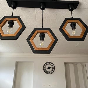 Peut inclure: Un luminaire suspendu moderne à trois lumières avec une structure en métal noir et des accents hexagonaux en bois. Chaque lumière est équipée d'une ampoule Edison visible. Le luminaire est monté sur un plafond blanc.