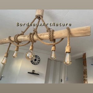 May include: A ceiling light fixture featuring a horizontal light-colored wooden beam suspended by thick jute rope. Five Edison-style light bulbs hang at varying lengths from the beam, each wrapped in rope. The text BordeauxArtNature is visible.