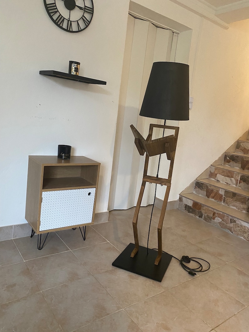 May include: A wooden floor lamp shaped like a person reading a book. The lamp has a black lampshade and a black base. The lamp is in a room with a white tiled floor and a white wall.