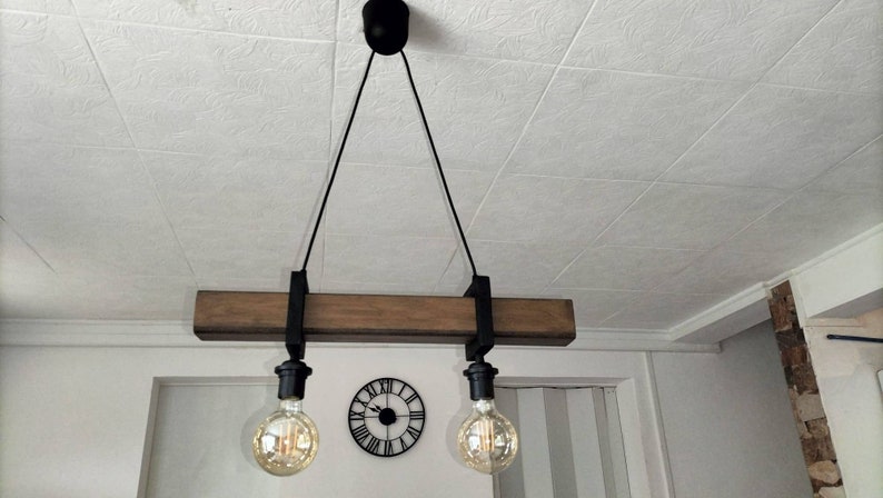 May include: A rustic wooden ceiling light fixture with two black metal hanging cords and Edison bulb light fixtures. The fixture is suspended from the ceiling with black cords.
