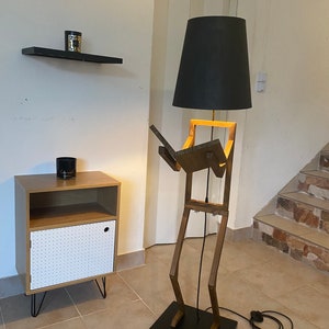 May include: A wooden floor lamp shaped like a person with a black lampshade. The lamp is standing on a black base and has a black cord.