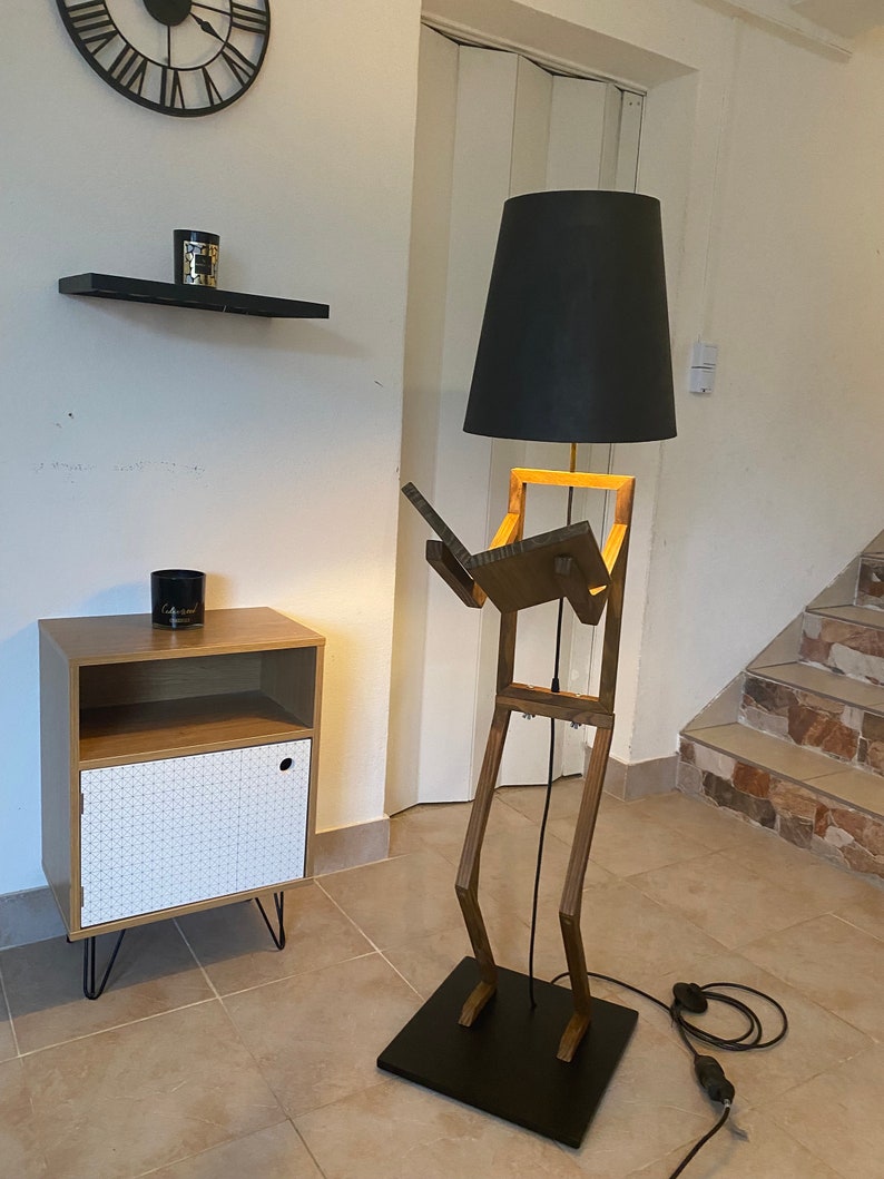 May include: A wooden floor lamp shaped like a person with a black lampshade. The lamp is standing on a black base and has a black cord.