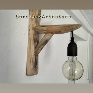 May include: A wall-mounted light fixture crafted from light-colored natural wood, featuring a vertical backplate, a horizontal shelf, and a diagonal support brace. A black socket hangs from the shelf holding a clear, round glass light bulb. The text BordeauxArtNature is visible.