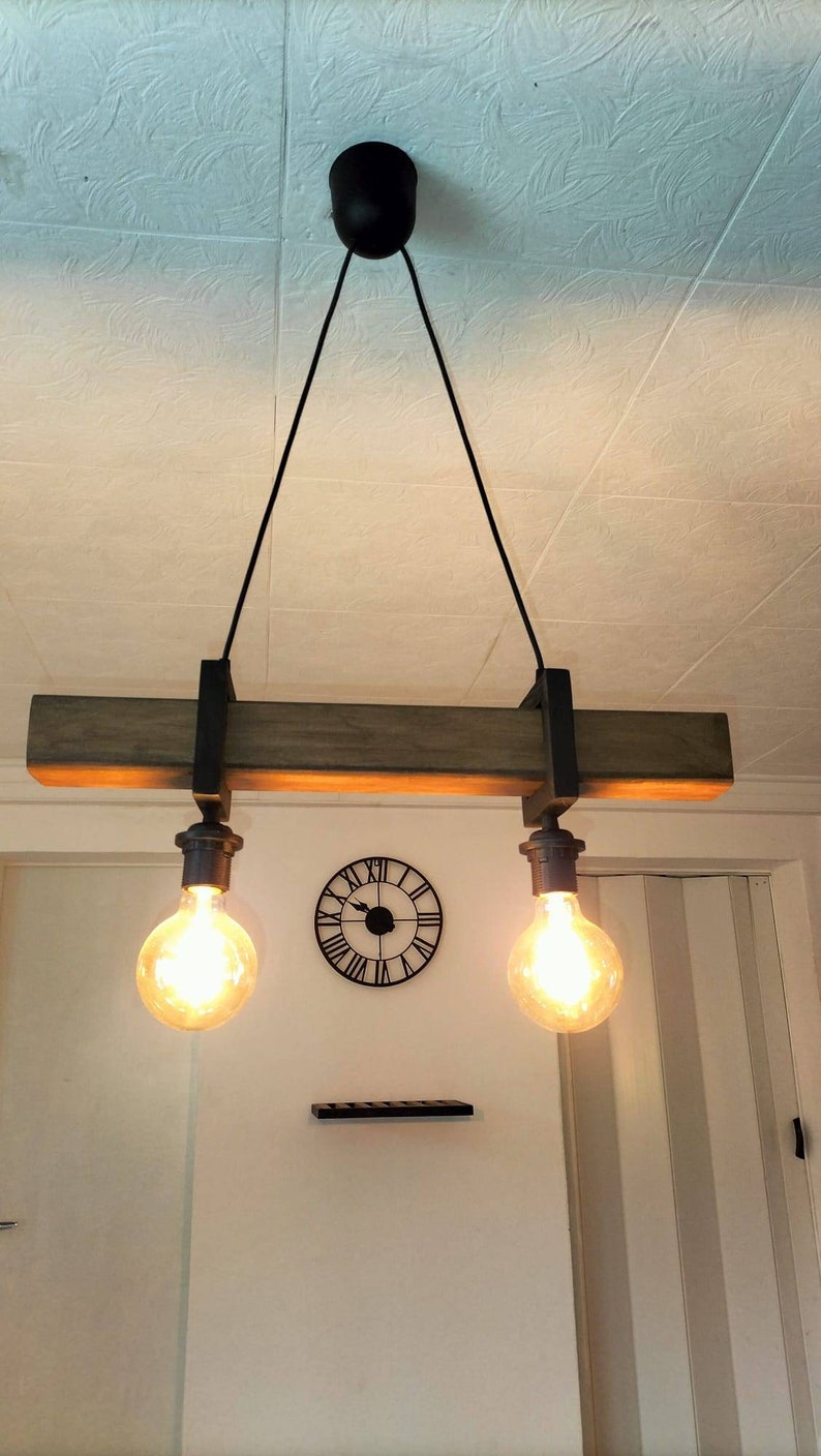 May include: A rustic wooden beam pendant light fixture with two exposed light bulbs. The fixture is suspended from the ceiling by black cords and has a black metal frame.