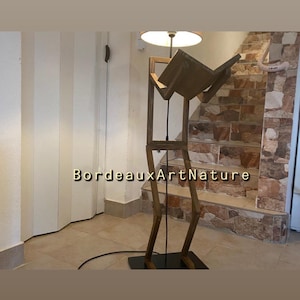 Può includere: Una lampada da terra realizzata in legno marrone scuro, caratterizzata da un design a figura umanoide con un supporto a forma di libro sulla parte superiore e un paralume circolare. Il testo BordeauxArtNature è visibile al centro della struttura.