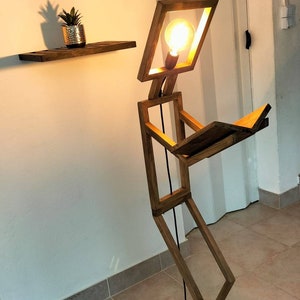 Peut inclure: Une lampe en bois en forme de personne lisant un livre. La lampe a une ampoule dans la t&ecirc;te et une base noire.