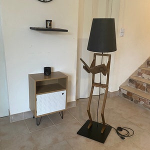 May include: A wooden floor lamp shaped like a person with a black lampshade. The lamp is standing next to a small wooden cabinet with a white door.