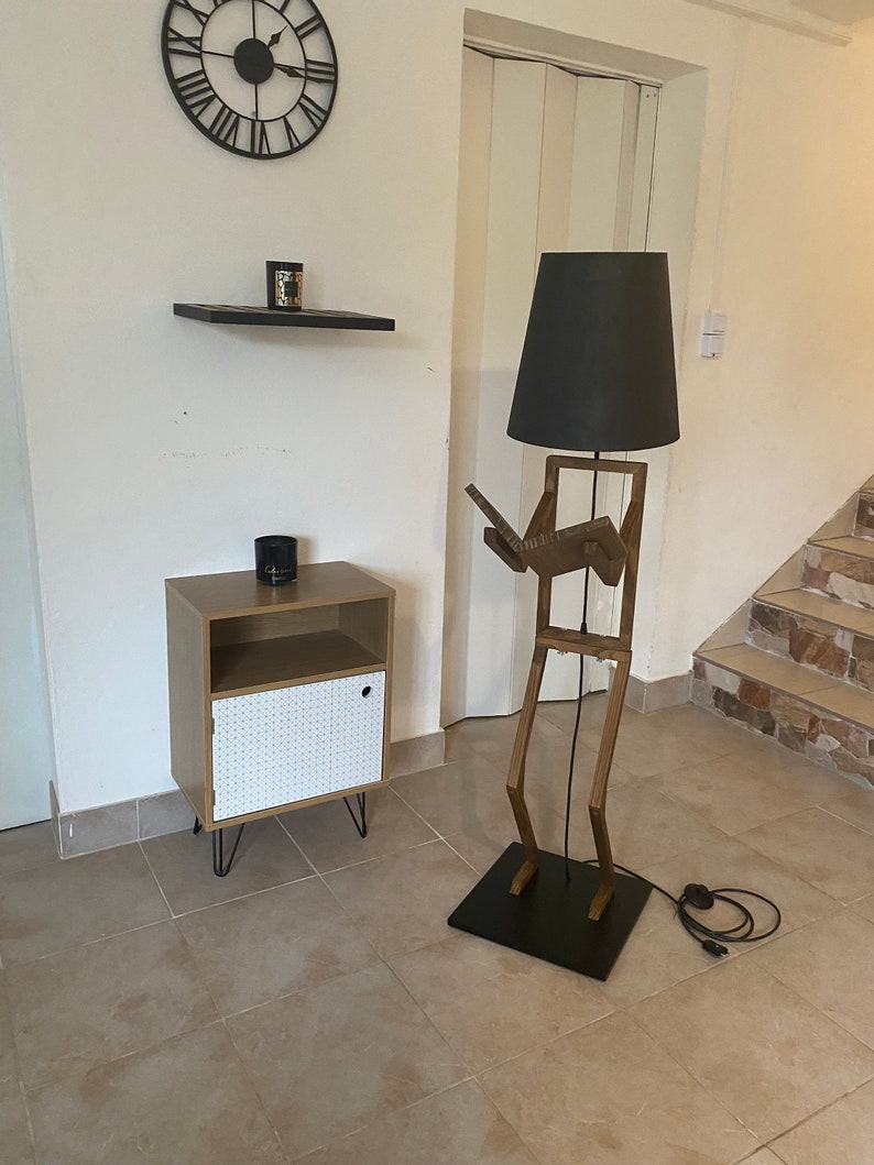 May include: A wooden floor lamp shaped like a person with a black lampshade. The lamp is standing next to a small wooden cabinet with a white door.