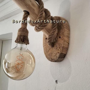May include: A wall-mounted light fixture featuring a natural wood log base and a circular wood backplate. A thick jute rope wraps around the log and suspends a large, clear glass globe bulb with a visible filament. The text BordeauxArtNature is visible.