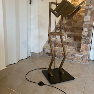 May include: A wooden floor lamp shaped like a person holding a book. The lamp has a black base and a black cord with a switch.