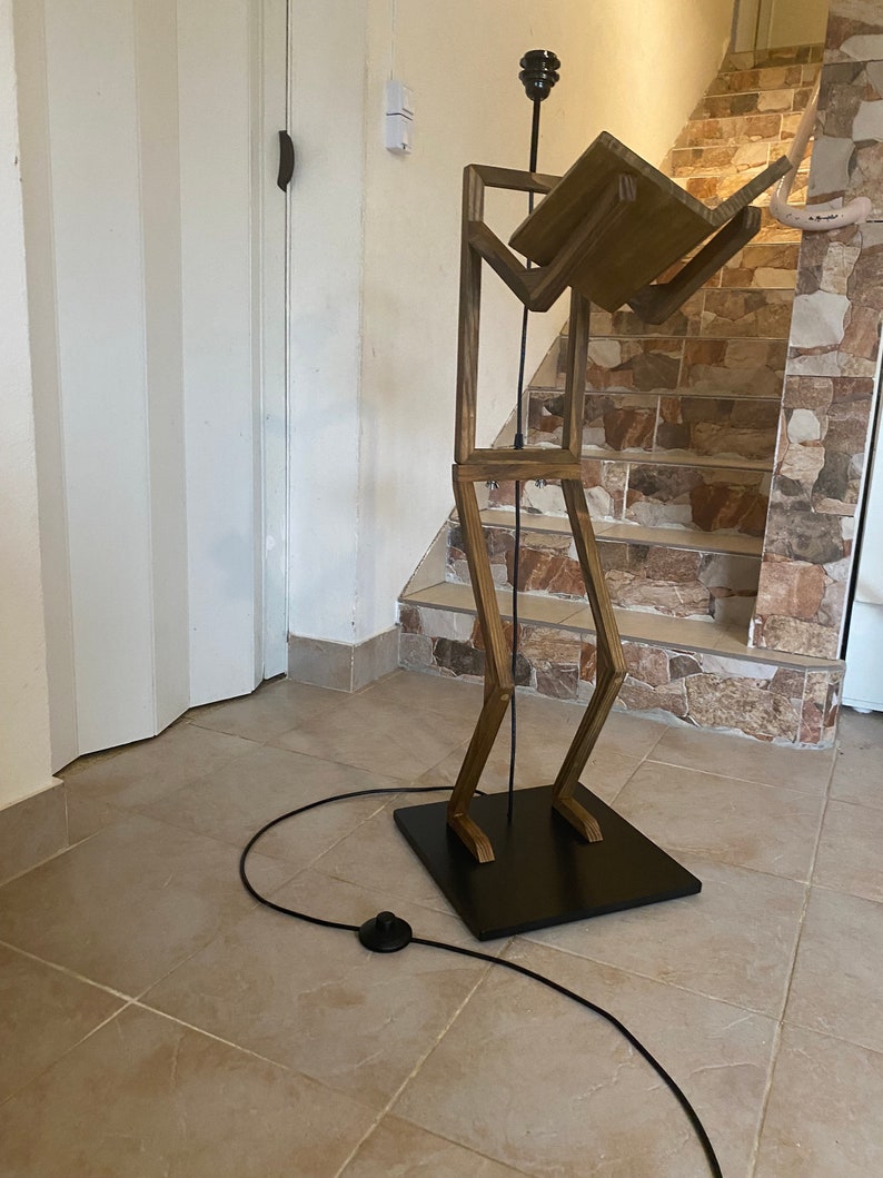 May include: A wooden floor lamp shaped like a person holding a book. The lamp has a black base and a black cord with a switch.