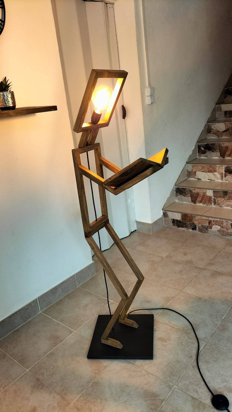 Peut inclure: Un lampadaire en bois en forme de personne lisant un livre. La lampe a une ampoule dans la t&ecirc;te et un cordon noir.
