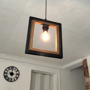 May include: A square pendant light fixture with a black metal frame and a wood accent. The light fixture has a single light bulb inside.