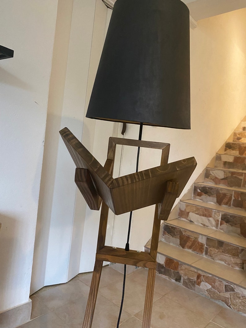 May include: A wooden floor lamp with a black lampshade. The lamp base is made of wood and has a unique design that resembles an open book. The lamp is standing on a tiled floor.