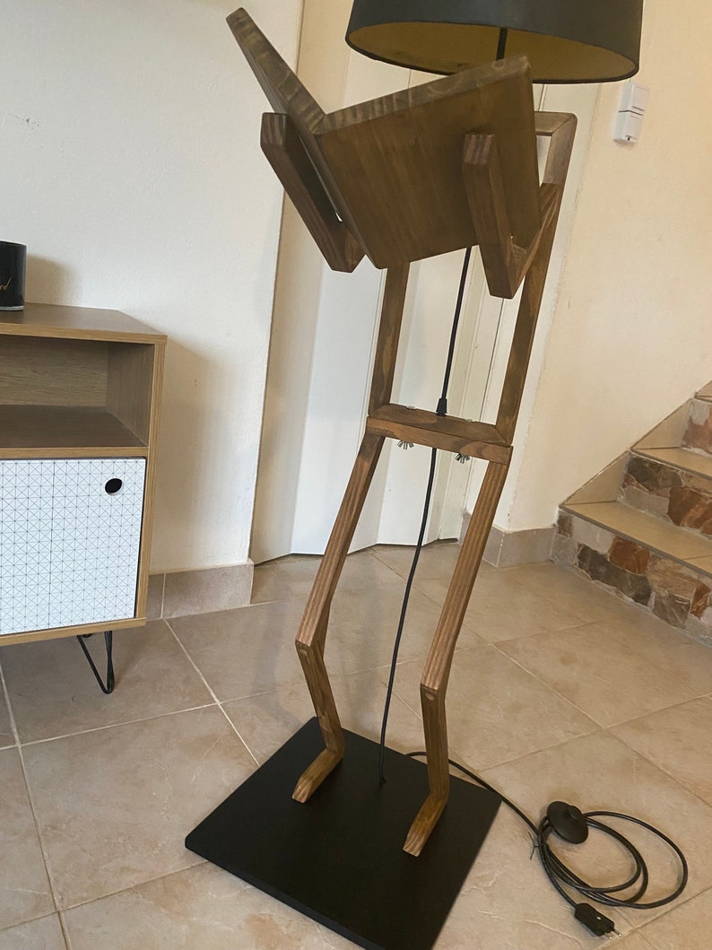 May include: A wooden floor lamp with a black shade and a wooden base shaped like a person. The lamp has a black cord and a black plug.