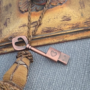 May include: A copper-colored USB flash drive shaped like a key, featuring a heart-shaped cutout in the key head and a heart detail on the USB connector. The key is attached to a decorative tassel and a rope-like cord.