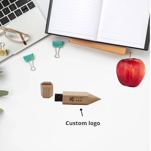 May include: A wooden pencil-shaped USB drive with a custom logo and the text "TEXAS A&M" engraved on the side. The drive is next to a red apple and a white surface.