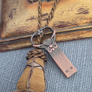 May include: A copper-colored USB flash drive with a decorative heart-shaped loop and a braided cord. The loop features floral and heart accents. The background includes a gold-toned, ornate surface and a tassel.