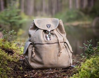 Puede incluir: Una mochila de lona color beige con cierre de solapa y cordón. La mochila tiene dos bolsillos laterales y un pequeño emblema en la solapa. Está sobre un lecho de musgo y hojas en un entorno forestal.