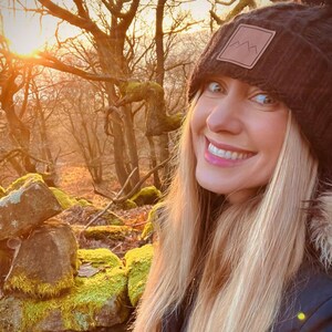 Peut inclure: Une femme aux longs cheveux blonds sourit &agrave; la cam&eacute;ra tout en portant un bonnet en tricot noir avec un patch brun repr&eacute;sentant une cha&icirc;ne de montagnes. Elle se tient dans un d&eacute;cor forestier avec un mur de pierre recouvert de mousse derri&egrave;re elle.
