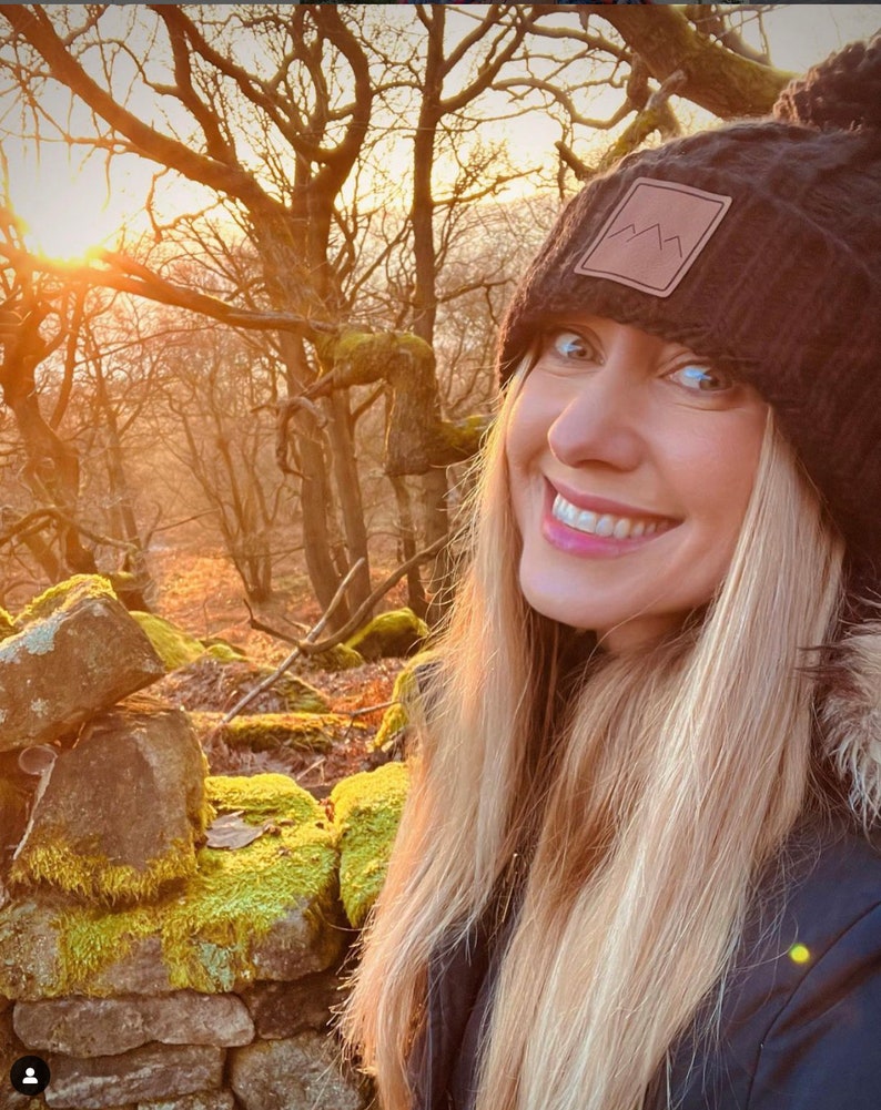 Peut inclure: Une femme aux longs cheveux blonds sourit &agrave; la cam&eacute;ra tout en portant un bonnet en tricot noir avec un patch brun repr&eacute;sentant une cha&icirc;ne de montagnes. Elle se tient dans un d&eacute;cor forestier avec un mur de pierre recouvert de mousse derri&egrave;re elle.