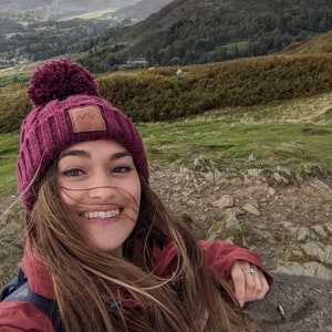 May include: A woman wearing a burgundy knit beanie with a brown patch and a burgundy jacket smiles at the camera. The beanie has a large pompom on top. The woman is standing in a mountainous landscape.