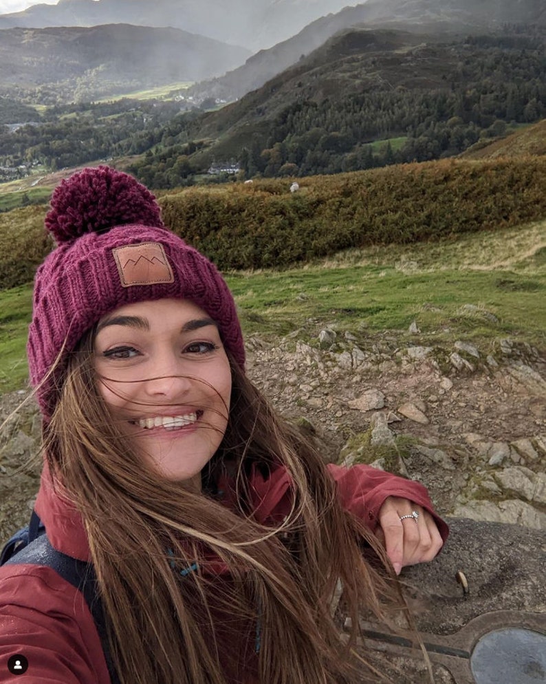 May include: A woman wearing a burgundy knit beanie with a brown patch and a burgundy jacket smiles at the camera. The beanie has a large pompom on top. The woman is standing in a mountainous landscape.
