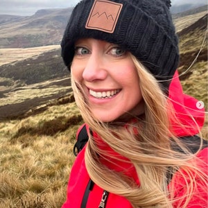 Peut inclure: Une femme portant une veste rouge et un bonnet noir en tricot avec un patch en cuir brun repr&eacute;sentant un logo de montagne. Ses cheveux sont blonds et flottent au vent. Elle se tient dans un champ d'herbe haute.