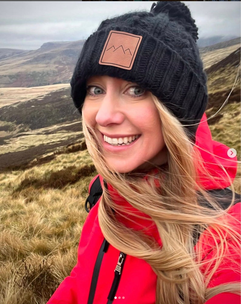 Peut inclure: Une femme portant une veste rouge et un bonnet noir en tricot avec un patch en cuir brun repr&eacute;sentant un logo de montagne. Ses cheveux sont blonds et flottent au vent. Elle se tient dans un champ d'herbe haute.