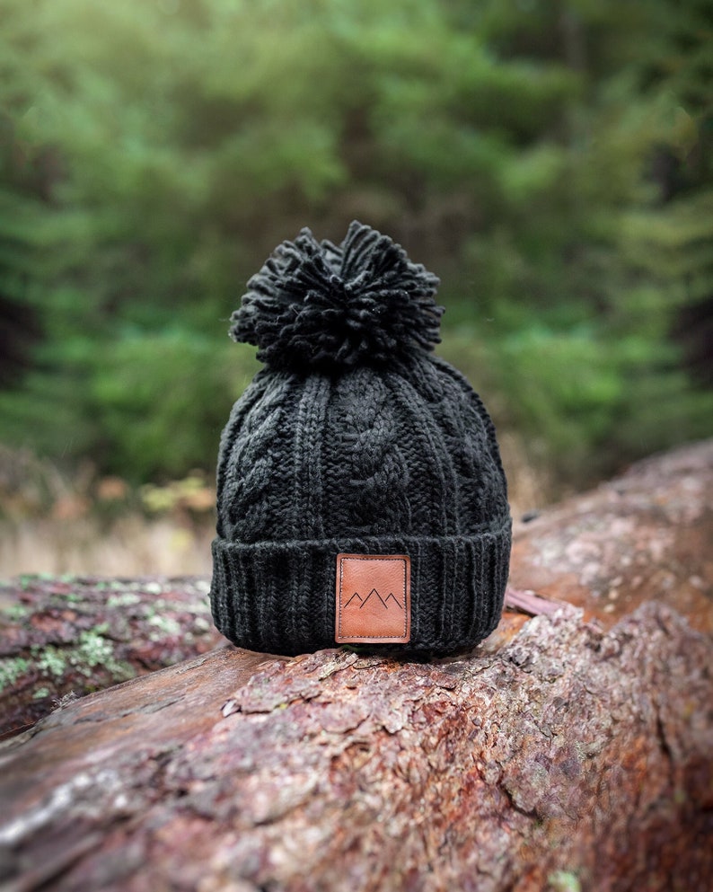 Peut inclure: Bonnet en tricot noir avec un patch en cuir marron orn&eacute; d'un logo de montagne. Le bonnet a un pompon moelleux sur le dessus.