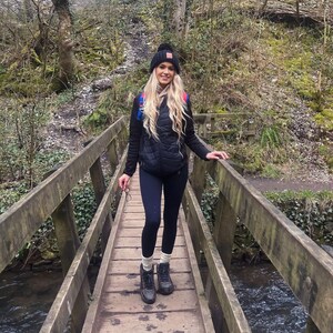 May include: A woman wearing a black puffer vest, black leggings, and black hiking boots stands on a wooden bridge over a stream. She is wearing a black beanie with a red and white logo.