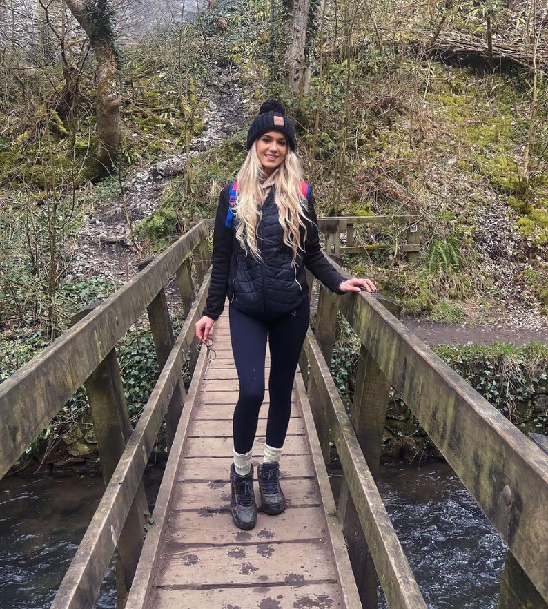 May include: A woman wearing a black puffer vest, black leggings, and black hiking boots stands on a wooden bridge over a stream. She is wearing a black beanie with a red and white logo.