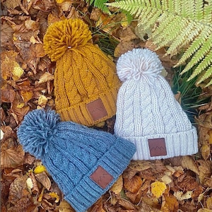 May include: Three knitted winter hats with pom poms. The hats are in different colors: mustard yellow, light pink, and blue. Each hat has a brown leather patch with a mountain logo.