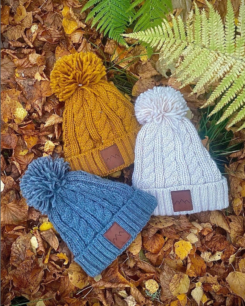 May include: Three knitted winter hats with pom poms. The hats are in different colors: mustard yellow, light pink, and blue. Each hat has a brown leather patch with a mountain logo.
