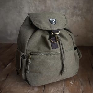 Może przedstawiać: Zielony plecak z płótna z sznurkiem do ściągania i małą kieszenią z przodu. Plecak ma czarno-białe logo na przedniej klapie.