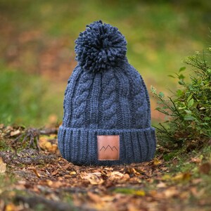 May include: A dark blue knitted beanie with a pom pom on top and a brown leather patch with a mountain logo. The beanie is sitting on a bed of fallen leaves.