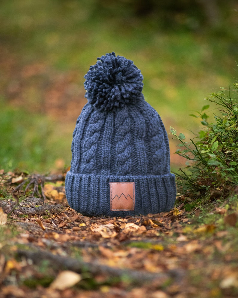 May include: A dark blue knitted beanie with a pom pom on top and a brown leather patch with a mountain logo. The beanie is sitting on a bed of fallen leaves.