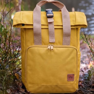 Puede incluir: Mochila de color amarillo mostaza con cierre enrollable y un bolsillo frontal con cremallera. La bolsa tiene correas color canela y un parche de cuero marrón con un diseño de montaña. La mochila está sobre un fondo borroso al aire libre.