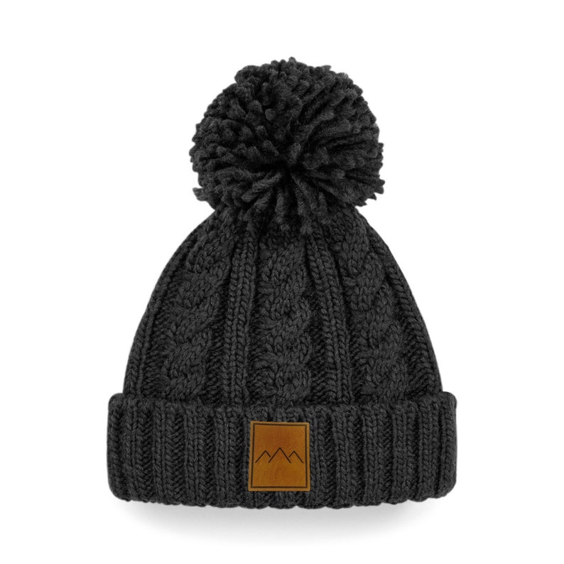 Peut inclure: Un bonnet en tricot noir avec un gros pompon sur le dessus et un patch en cuir marron avec un motif de montagne sur le devant.