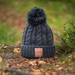 Peut inclure: Un bonnet en tricot gris fonc&eacute; avec un pompon sur le dessus et un patch en cuir marron avec un logo de montagne. Le bonnet est pos&eacute; sur un lit de feuilles mortes.