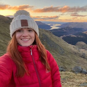 May include: A woman wearing a red puffer jacket and a white knit beanie with a pom-pom on top. She is standing on a mountaintop with a view of a valley and a lake in the distance.