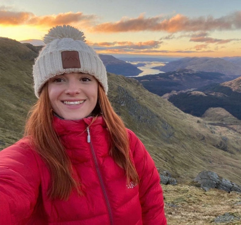 May include: A woman wearing a red puffer jacket and a white knit beanie with a pom-pom on top. She is standing on a mountaintop with a view of a valley and a lake in the distance.
