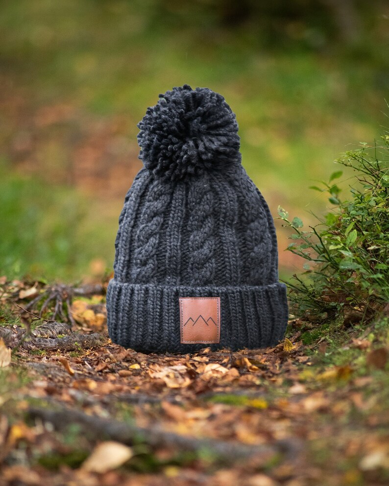 May include: A dark gray knitted beanie with a pom pom on top and a brown leather patch with a mountain logo. The beanie is sitting on a bed of fallen leaves.