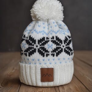 May include: A white knit beanie with a large white pom-pom on top. The hat features a pattern of black and light blue snowflake-like designs. A brown leather patch with a mountain graphic is sewn on the brim.
