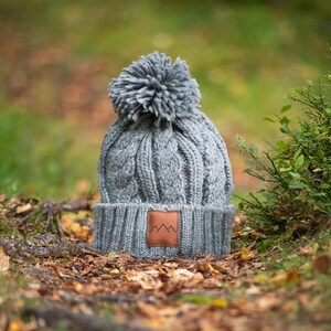 May include: Gray knitted beanie with a pom pom on top and a brown leather patch with a mountain logo. The beanie is sitting on a bed of fallen leaves.