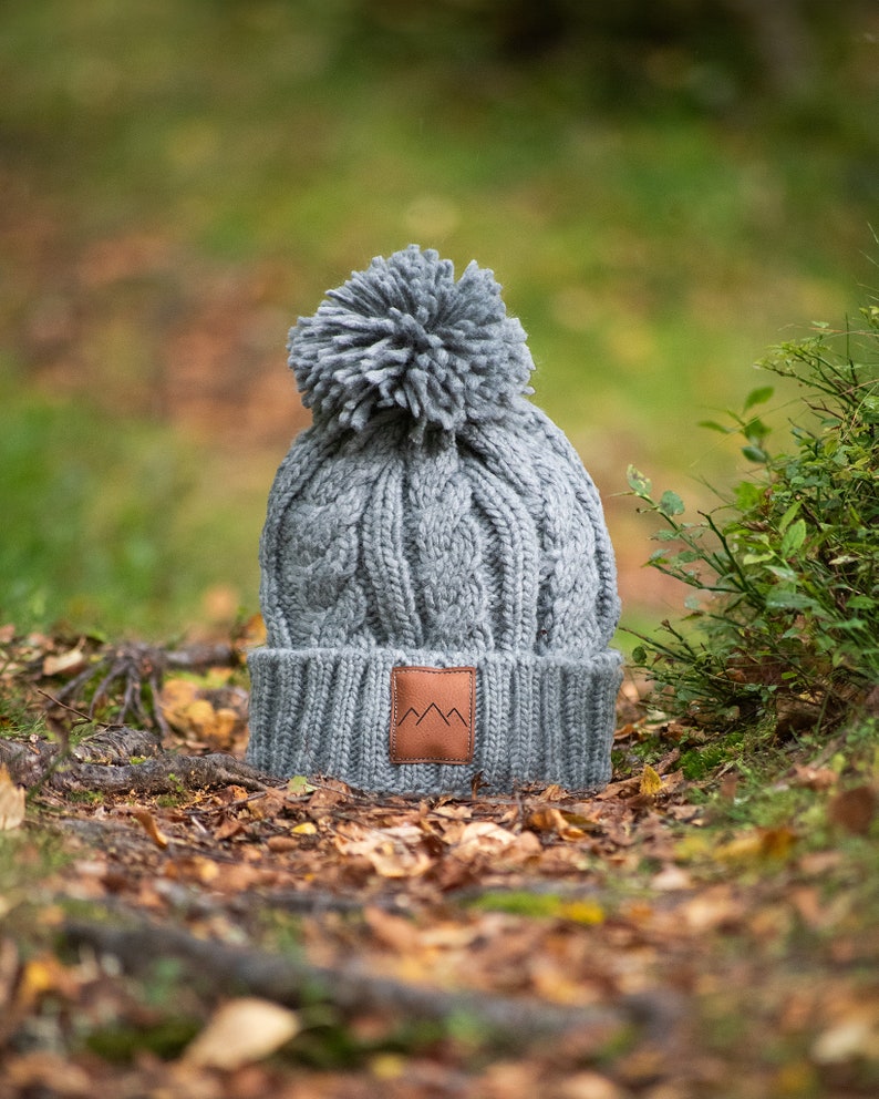 May include: Gray knitted beanie with a pom pom on top and a brown leather patch with a mountain logo. The beanie is sitting on a bed of fallen leaves.