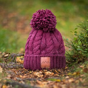 May include: A burgundy knitted beanie with a pom pom on top and a brown leather patch with a mountain logo. The beanie is sitting on a bed of fallen leaves.