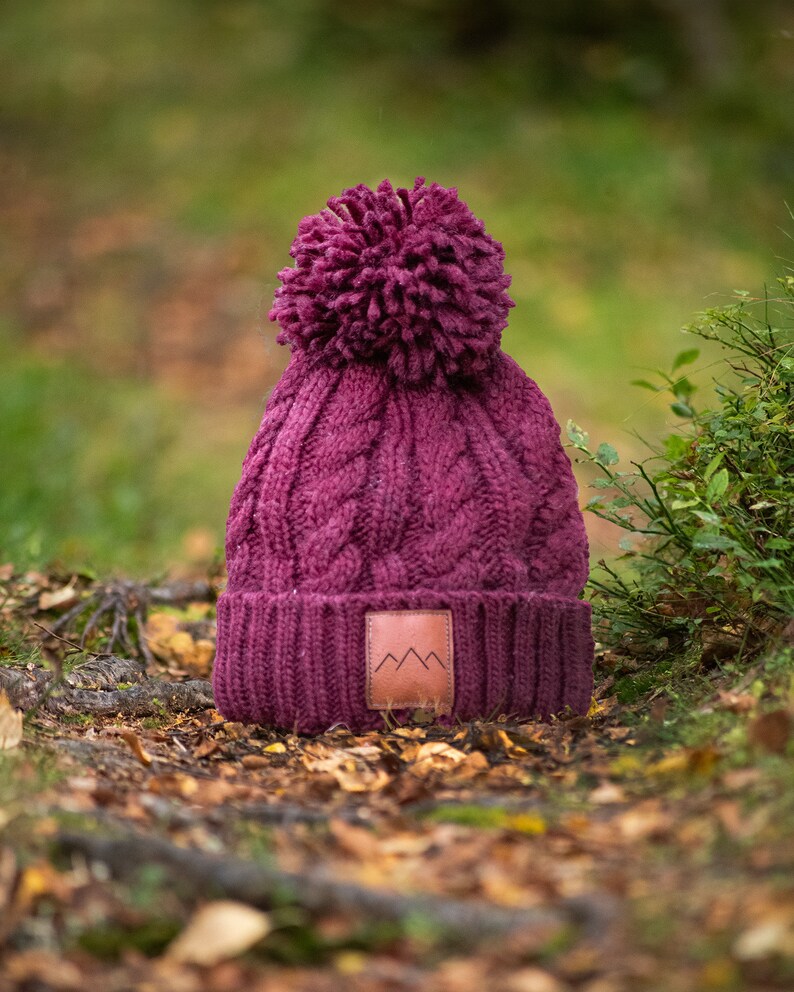 May include: A burgundy knitted beanie with a pom pom on top and a brown leather patch with a mountain logo. The beanie is sitting on a bed of fallen leaves.